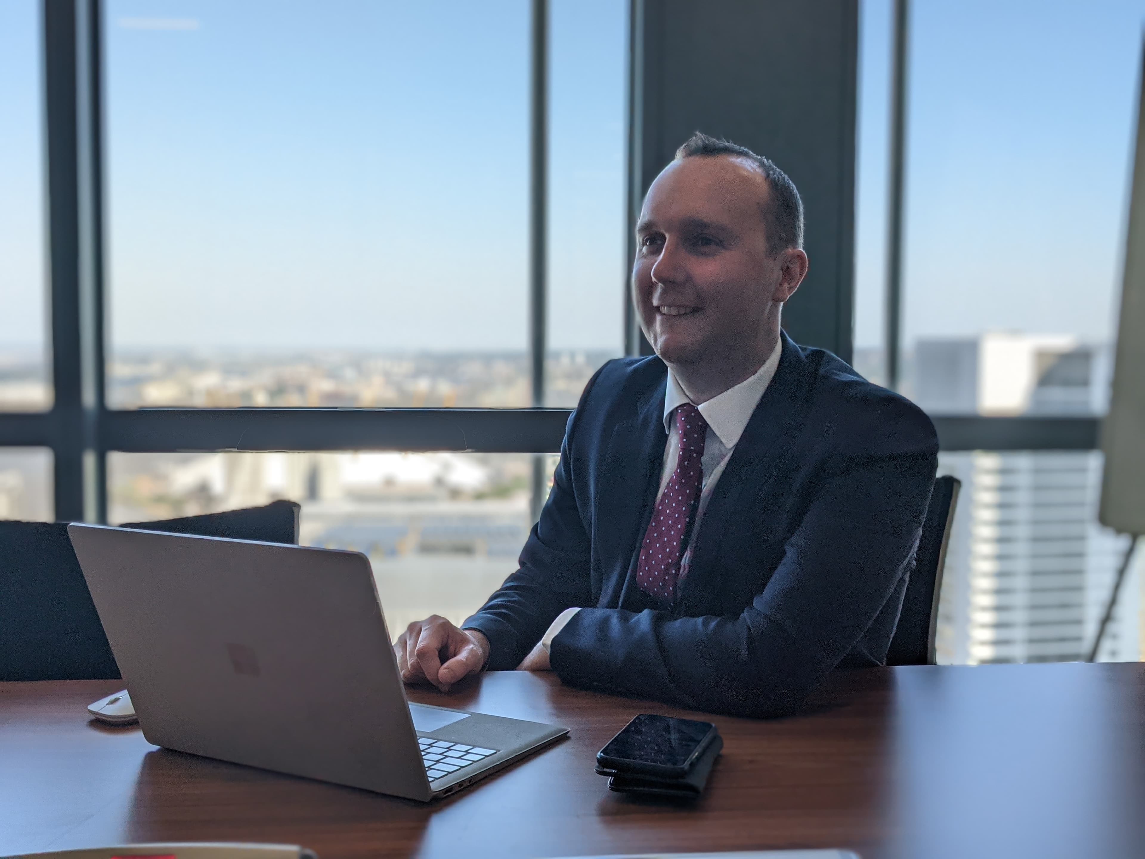 Smiling man in a suit at a desk with a laptop and city background.