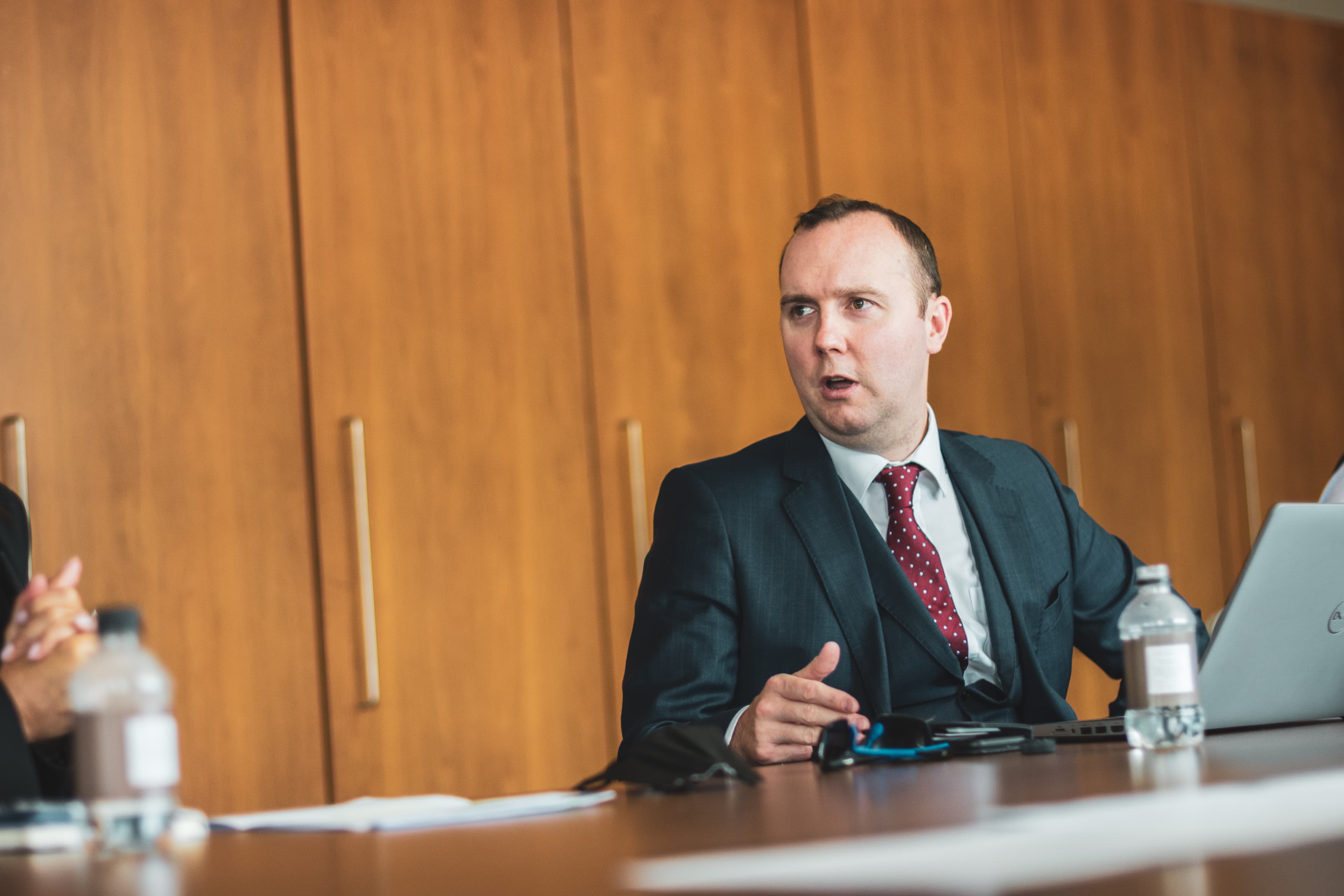 Man in a business suit and red tie gestures while speaking at a meeting table.